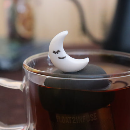 smiling white crescent moon tea infuser for loose leaf tea with a fine mesh stainless steel strainer that reads FLOAT2INFUSE  on a black base floating in a glass tea mug with tea kettle in the background