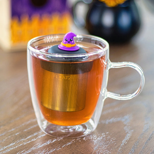 purple witch hat with a smiling face and an orange buckle tea infuser for loose leaf tea with a fine mesh stainless steel tea strainer that reads FLOAT2INFUSE on a black base floating in a glass tea mug with a cauldron in the background
