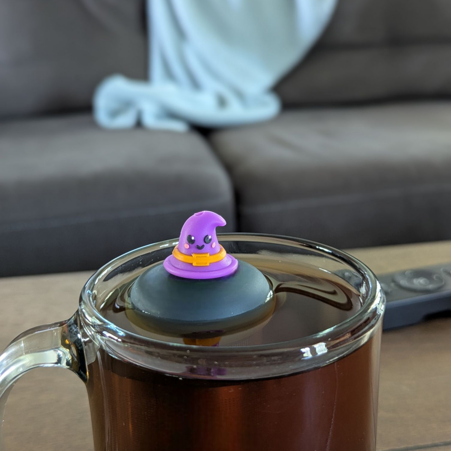 purple witch hat with a smiling face and an orange buckle tea infuser for loose leaf tea with a fine mesh stainless steel tea strainer that reads FLOAT2INFUSE on a black base floating in a glass tea mug with a couch and blanket in the background