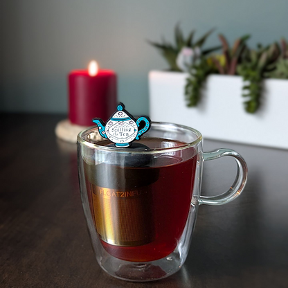blue and white tea kettle that reads "spilling the tea"  tea infuser for loose leaf tea with a fine mesh stainless steel tea infuser that reads FLOAT2INFUSE on a black base  floating in a glass tea mug with  plants and a candle in the background