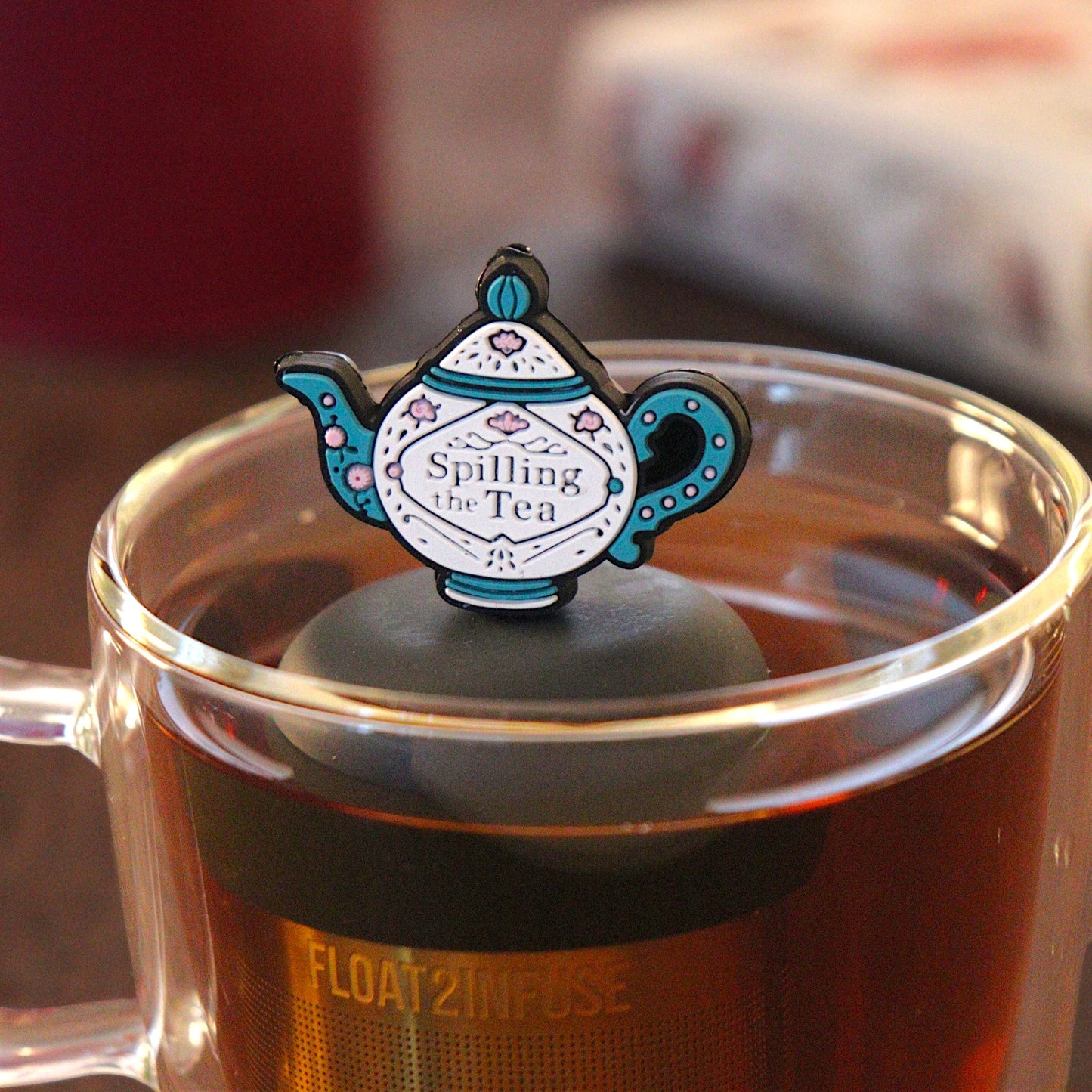blue and white tea kettle that reads "spilling the tea"  tea infuser for loose leaf tea with a fine mesh stainless steel tea infuser that reads FLOAT2INFUSE on a black base  floating in a glass tea mug with  loose leaf tea and books in the background