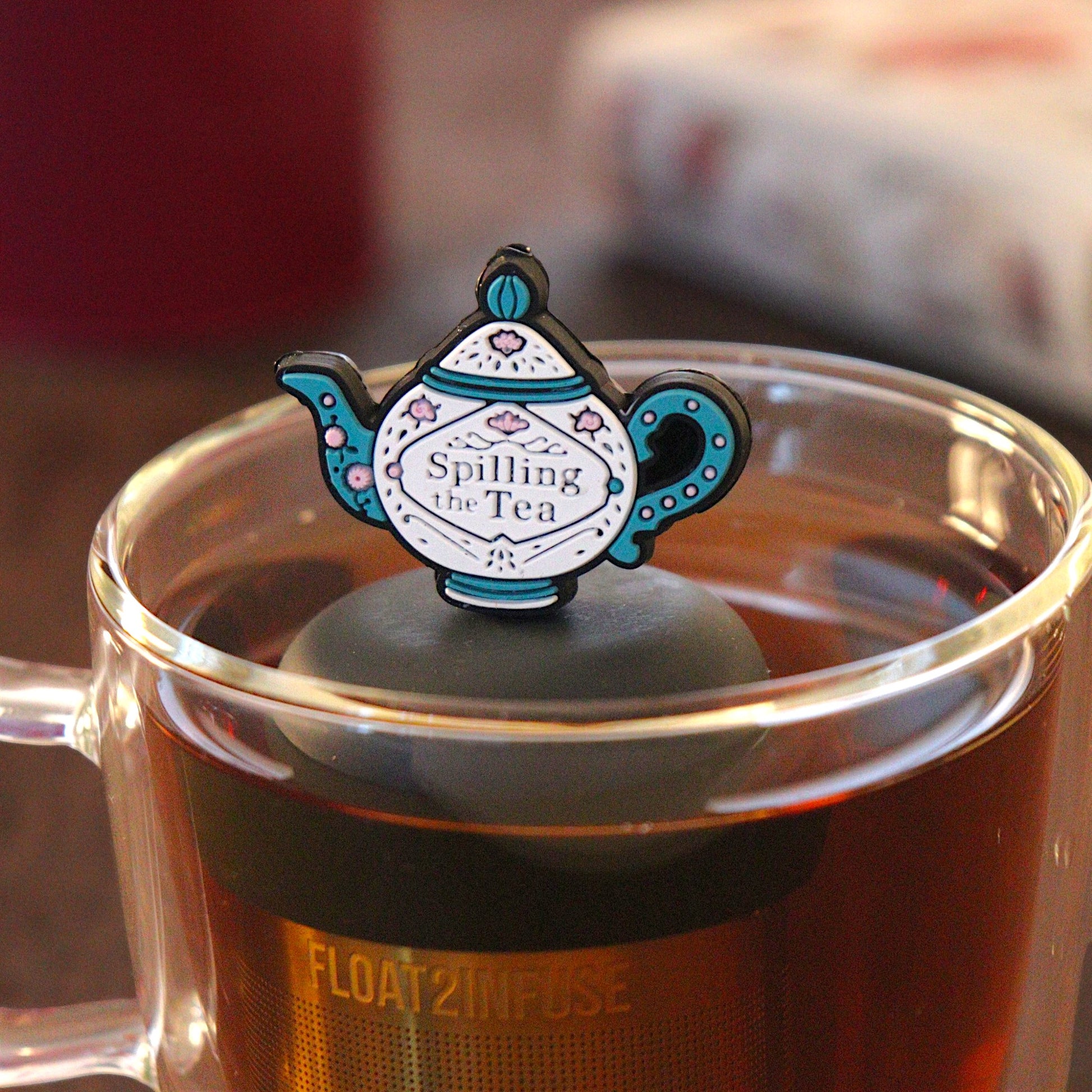 blue and white tea kettle that reads "spilling the tea"  tea infuser for loose leaf tea with a fine mesh stainless steel tea infuser that reads FLOAT2INFUSE on a black base  floating in a glass tea mug with  loose leaf tea and books in the background
