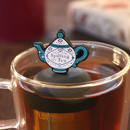 blue and white tea kettle that reads "spilling the tea"  tea infuser for loose leaf tea with a fine mesh stainless steel tea infuser that reads FLOAT2INFUSE on a black base  floating in a glass tea mug with  loose leaf tea and books in the background