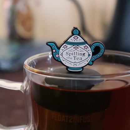 blue and white tea kettle that reads "spilling the tea" tea infuser for loose leaf tea  with a fine mesh stainless steel strainer that reads FLOAT2INFUSE on a black base floating in a glass tea mug with books in the background