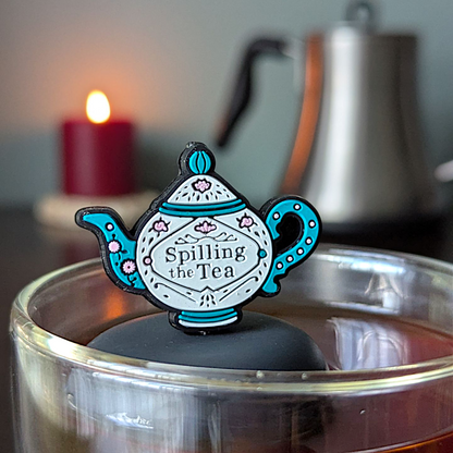 blue and white tea kettle that reads "spilling the tea"  tea infuser for loose leaf tea with a fine mesh stainless steel tea infuser that reads FLOAT2INFUSE on a black base  floating in a glass tea mug with candle and tea kettle in the background