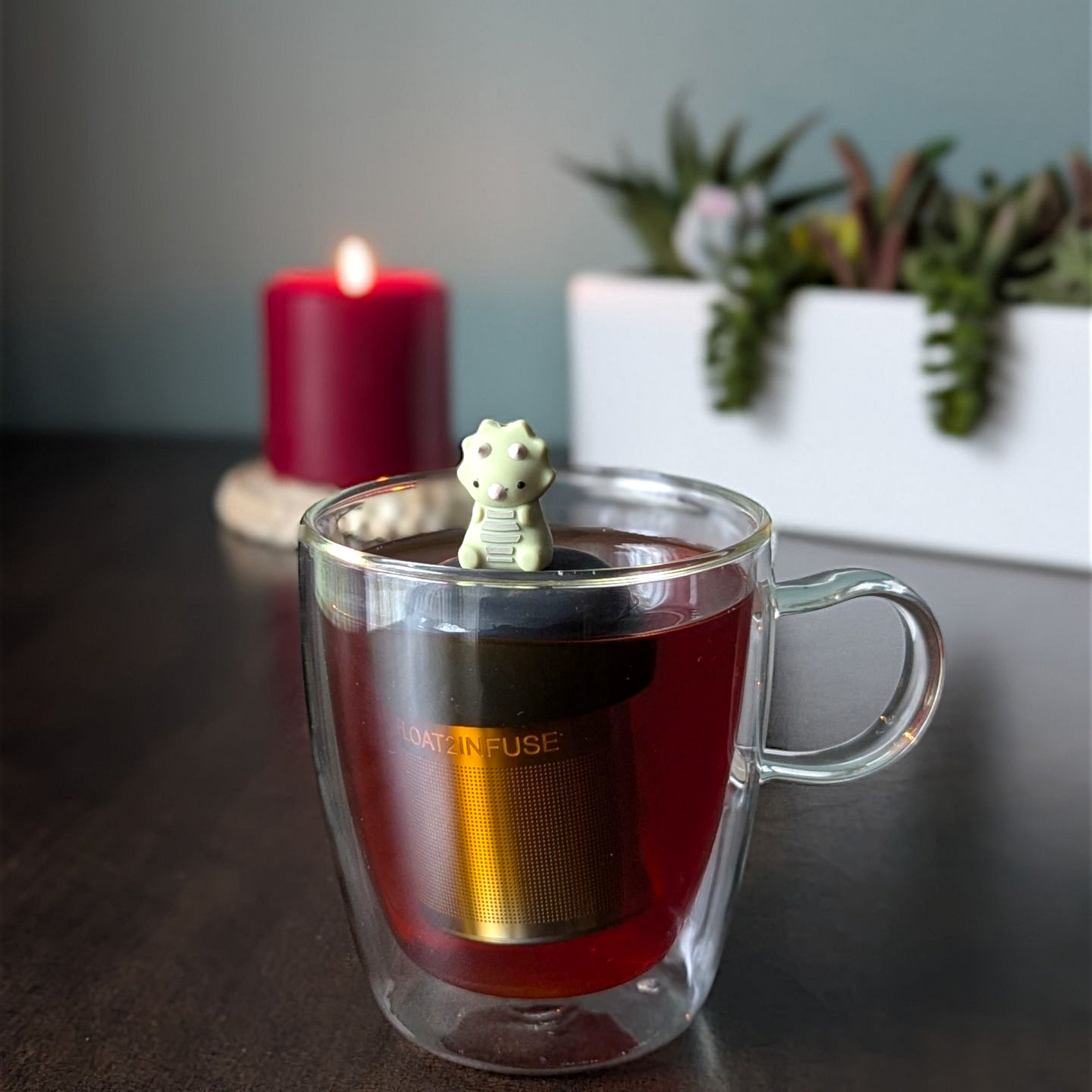 sage green triceratops dinosaur animal tea infuser for loose leaf tea  with a fine mesh stainless steel tea infuser that reads FLOAT2INFUSE on a black base floating in a glass tea mug with plants and a candle in the background
