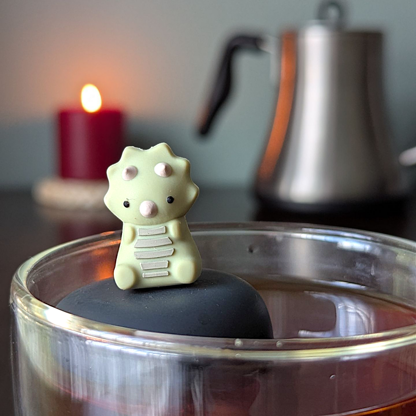 sage green triceratops dinosaur animal tea infuser for loose leaf tea  with a fine mesh stainless steel tea infuser that reads FLOAT2INFUSE on a black base floating in a glass tea mug with a tea kettle and a candle in the background