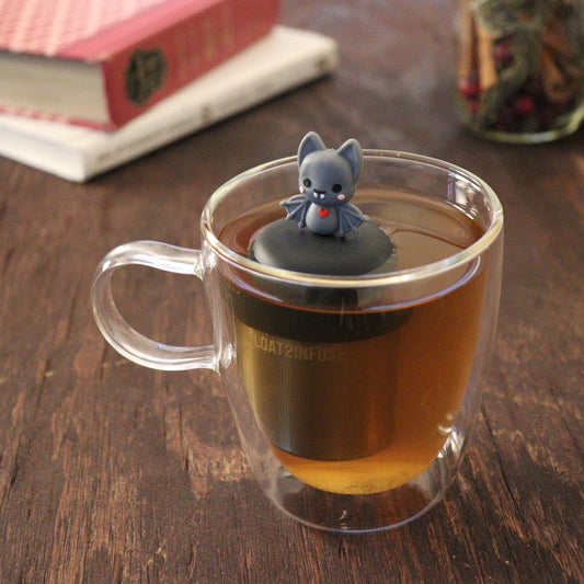 blushing grey vampire bat with red heart animal tea infuser for loose leaf tea  with a fine mesh stainless steel tea infuser that reads FLOAT2INFUSE on a black base floating in a glass tea mug with books and loose leaf tea in the background