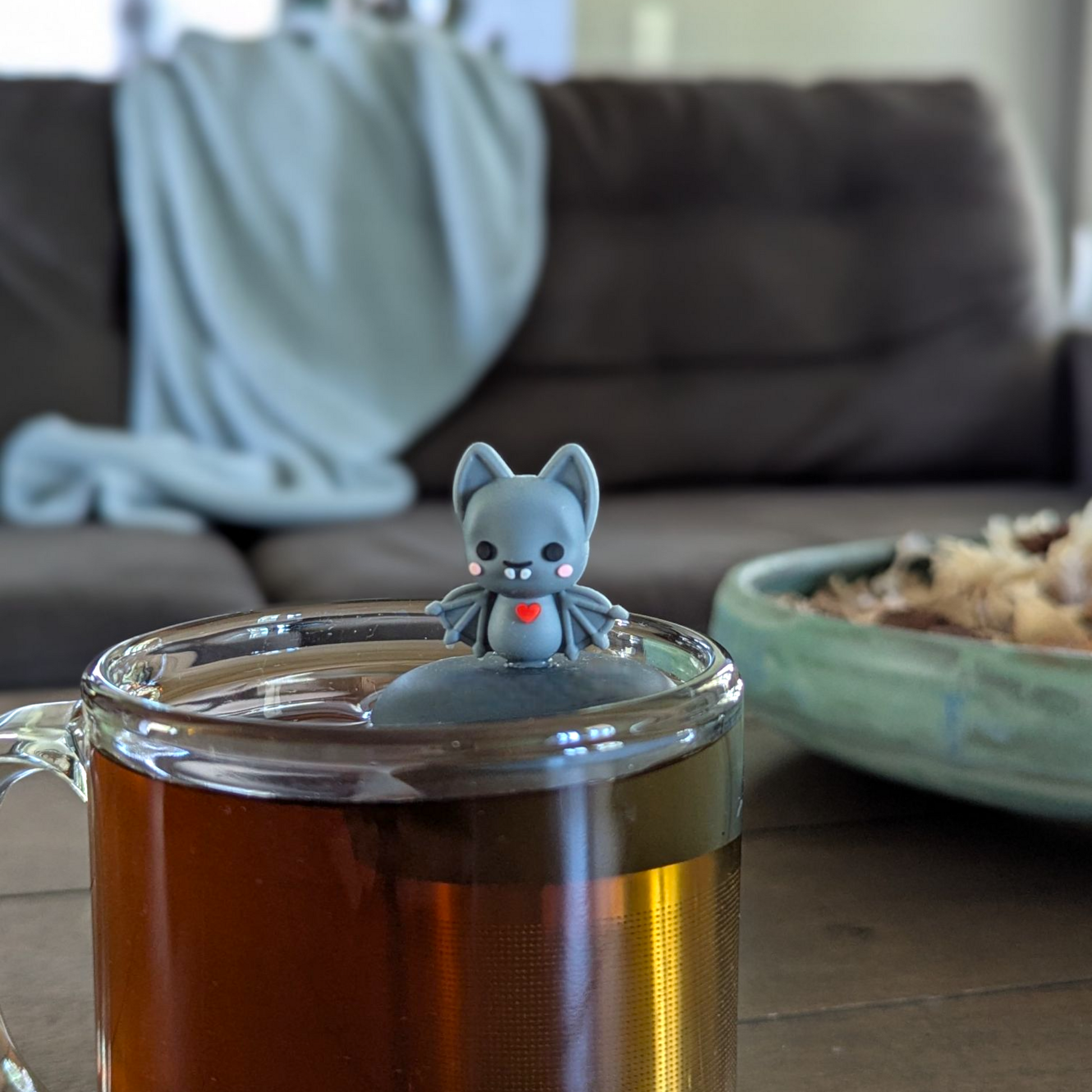 blushing grey vampire bat with red heart animal tea infuser for loose leaf tea with a fine mesh stainless steel tea infuser that reads FLOAT2INFUSE on a black base floating in a glass tea mug with a couch in the background