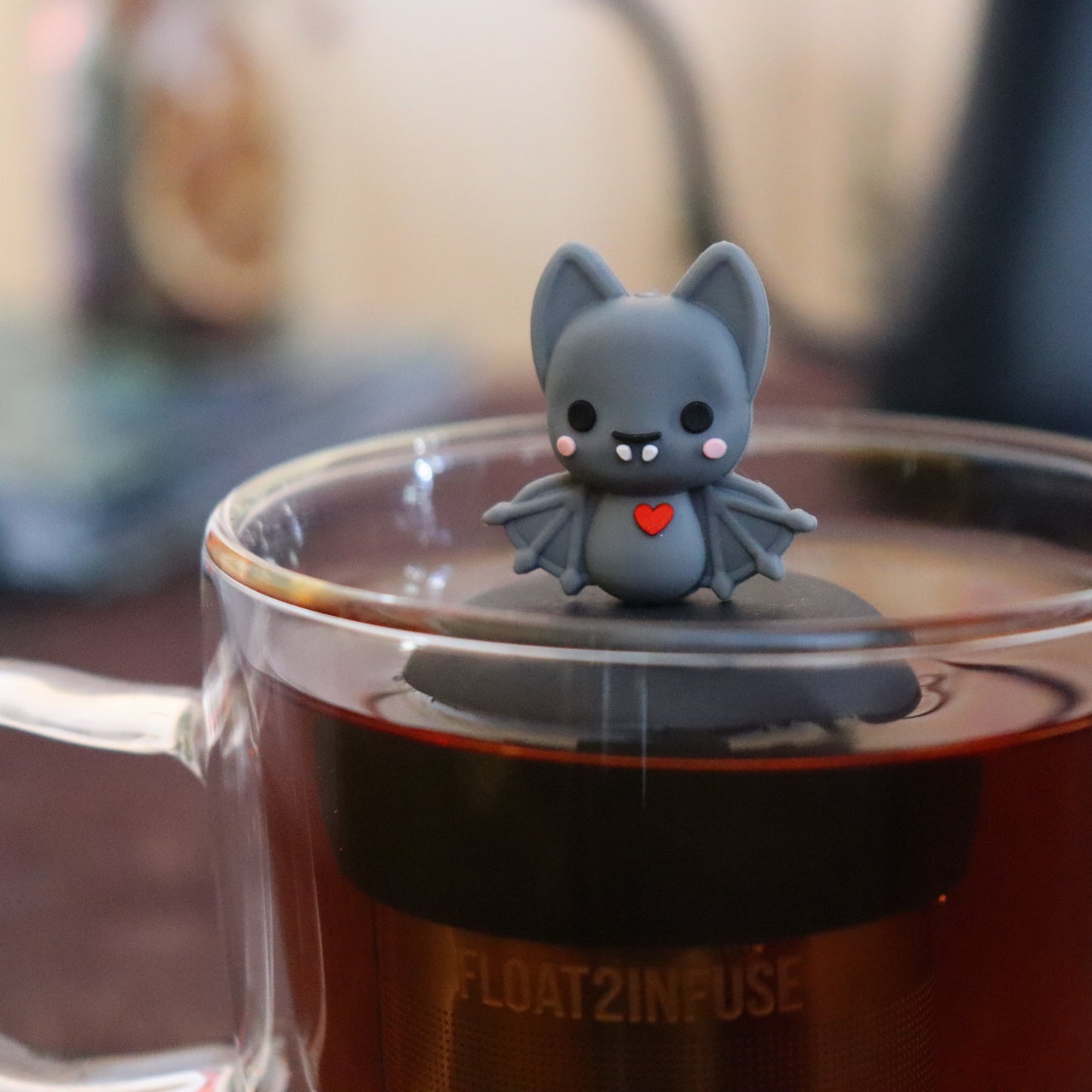 blushing grey vampire bat with red heart animal tea infuser for loose leaf tea with a fine mesh stainless steel tea infuser that reads FLOAT2INFUSE on a black base floating in a glass tea mug with tea kettle in the background