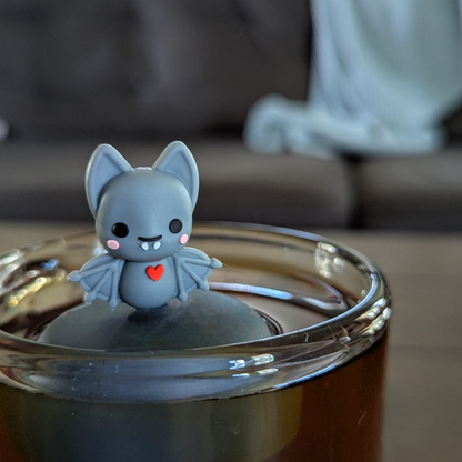 blushing grey vampire bat with red heart animal tea infuser for loose leaf tea  with a fine mesh stainless steel tea infuser that reads FLOAT2INFUSE on a black base floating in a glass tea mug with a couch in the background