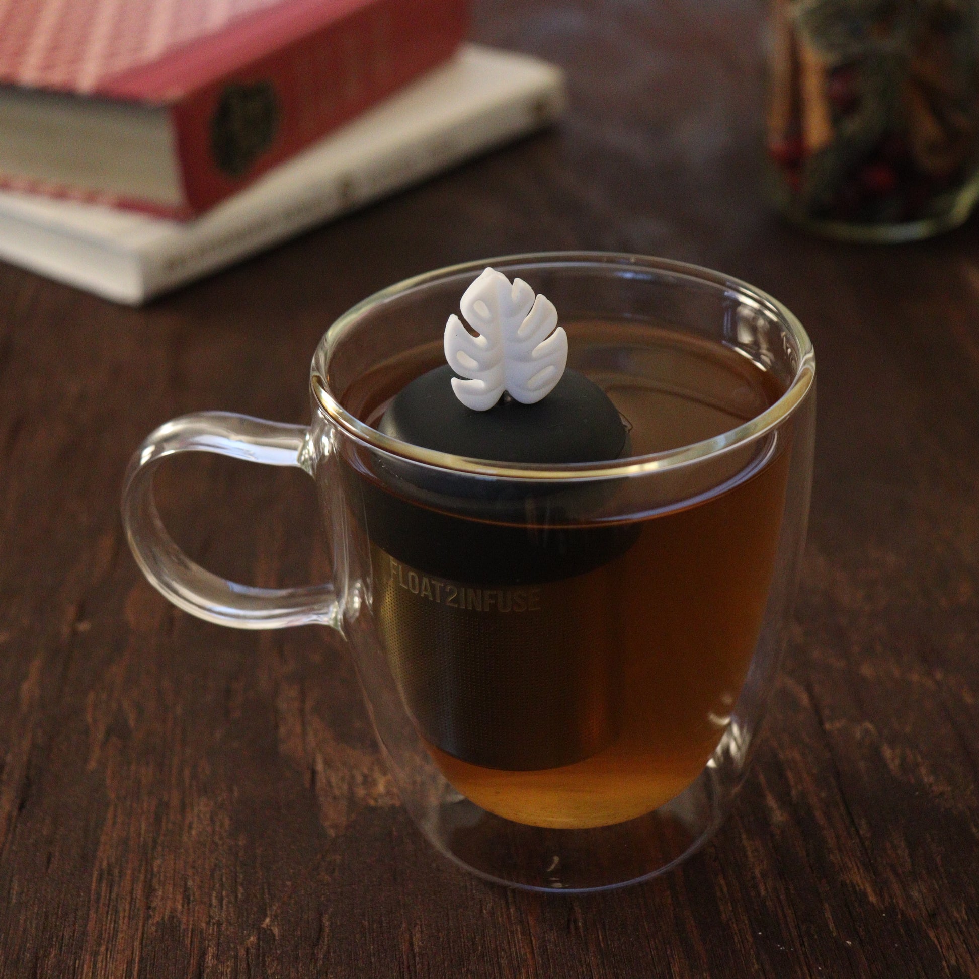 white monstera leaf tea infuser for loose leaf tea on a black base with a fine mesh stainless steel tea infuser that reads FLOAT2INFUSE floating in a glass tea mug with books and loose leaf tea in the background