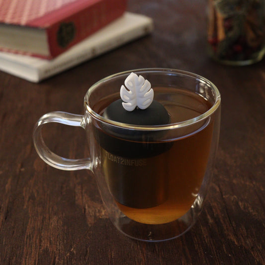 white monstera leaf tea infuser for loose leaf tea on a black base with a fine mesh stainless steel tea infuser that reads FLOAT2INFUSE floating in a glass tea mug with books and loose leaf tea in the background