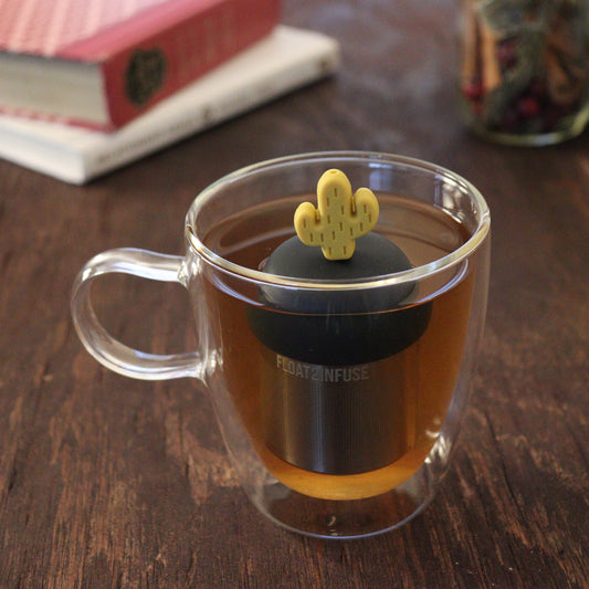 yellow cactus tea infuser for loose leaf tea  with a fine mesh stainless steel tea infuser that reads FLOAT2INFUSE on a black base floating in a glass tea mug with books and loose leaf tea in the background