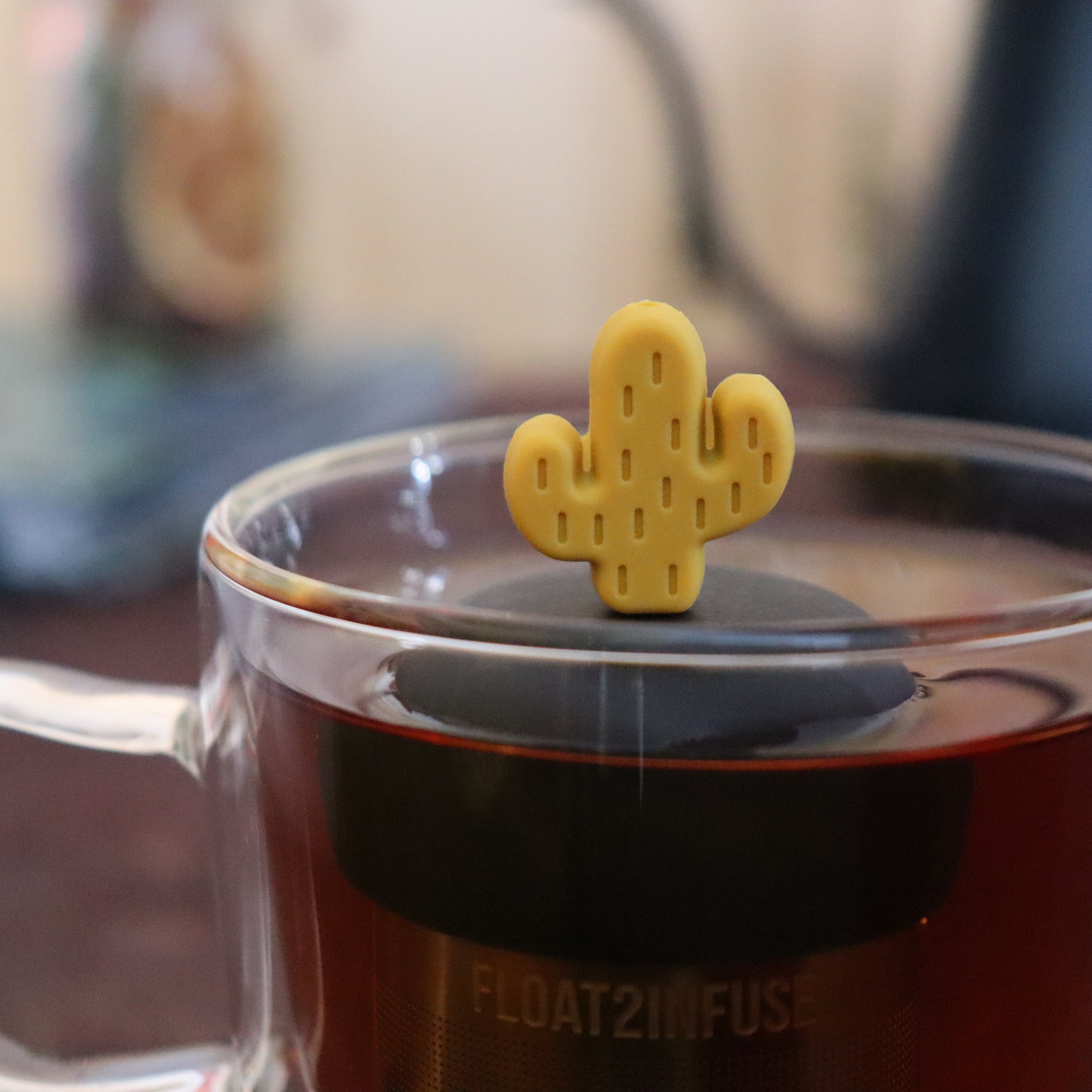 yellow cactus tea infuser for loose leaf tea  with a fine mesh stainless steel strainer that reads FLOAT2INFUSE on a black base floating in a glass tea mug with tea kettle in the background