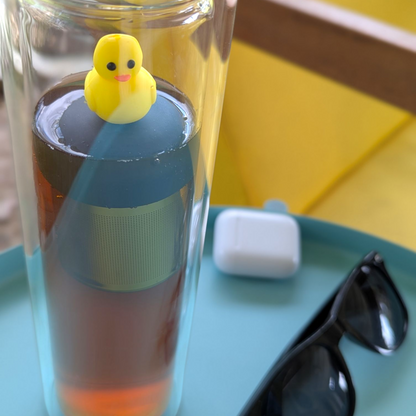 yellow rubber duck tea infuser for loose leaf tea with a fine mesh stainless steel tea strainer that reads FLOAT2INFUSE on a black base  floating in a glass travel bottle with sunglasses in the background