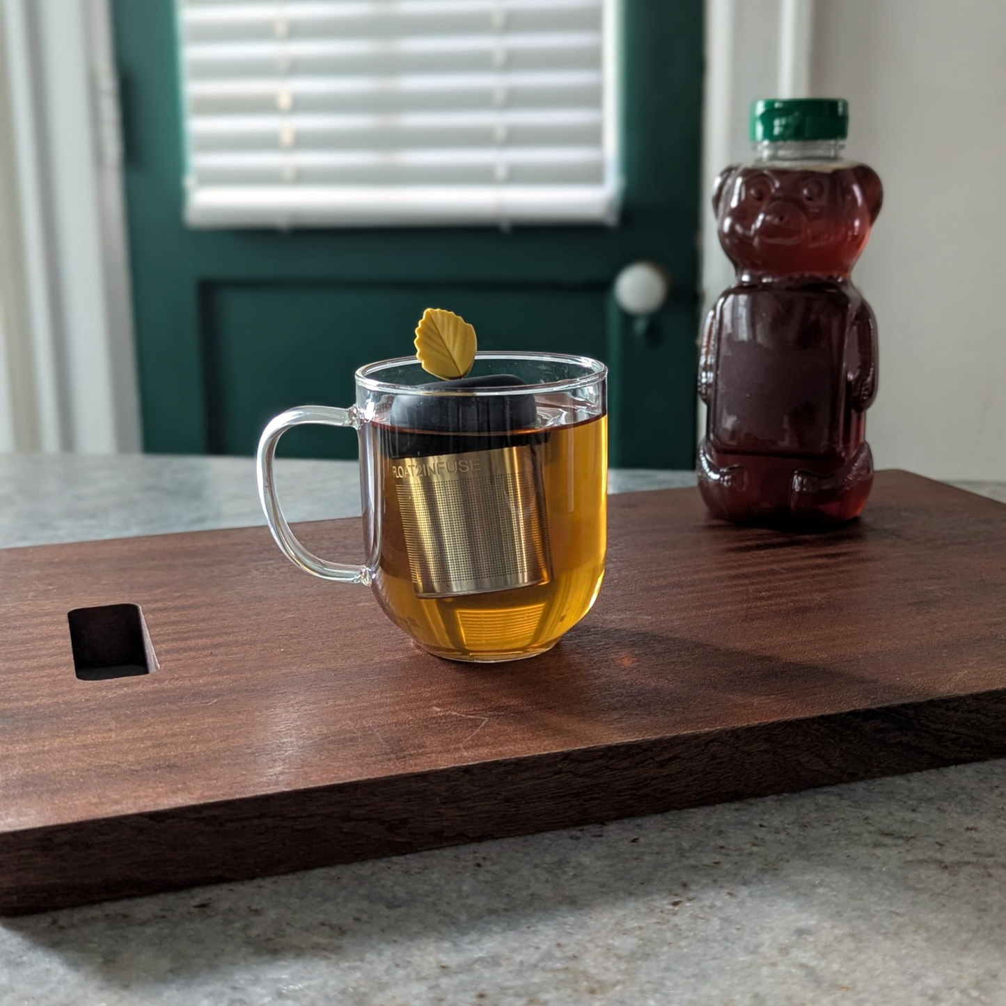 yellow fall leaf tea infuser for loose leaf tea  with a fine mesh stainless steel strainer that reads FLOAT2INFUSE on a black base floating in a glass tea mug with a honey bear in the background