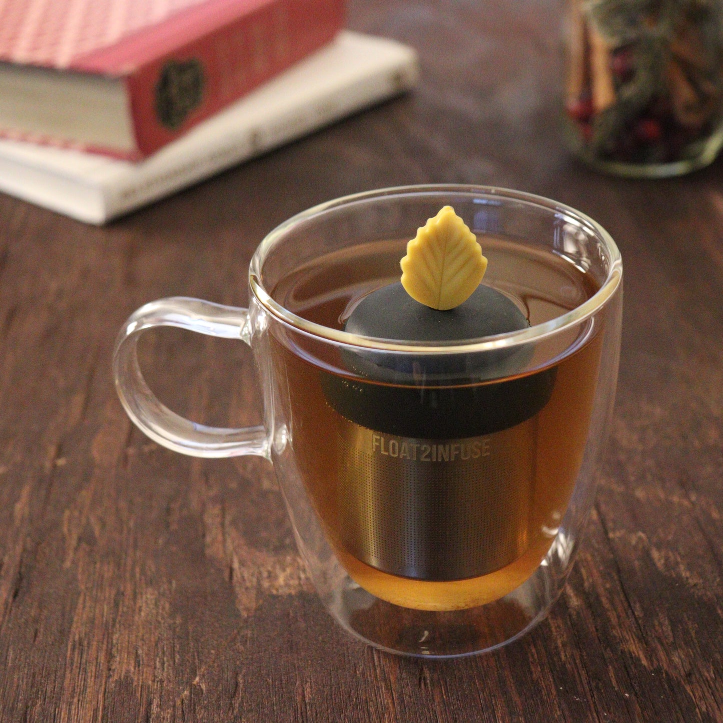 yellow fall leaf tea infuser for loose leaf tea  with a fine mesh stainless steel tea infuser that reads FLOAT2INFUSE on a black base floating in a glass tea mug with books and loose leaf tea in the background