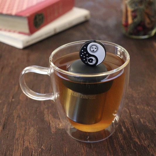 black and white yin yang with sun and moon tea infuser for loose leaf tea  with a fine mesh stainless steel tea infuser that reads FLOAT2INFUSE on a black base floating in a glass tea mug with books and loose leaf tea in the background