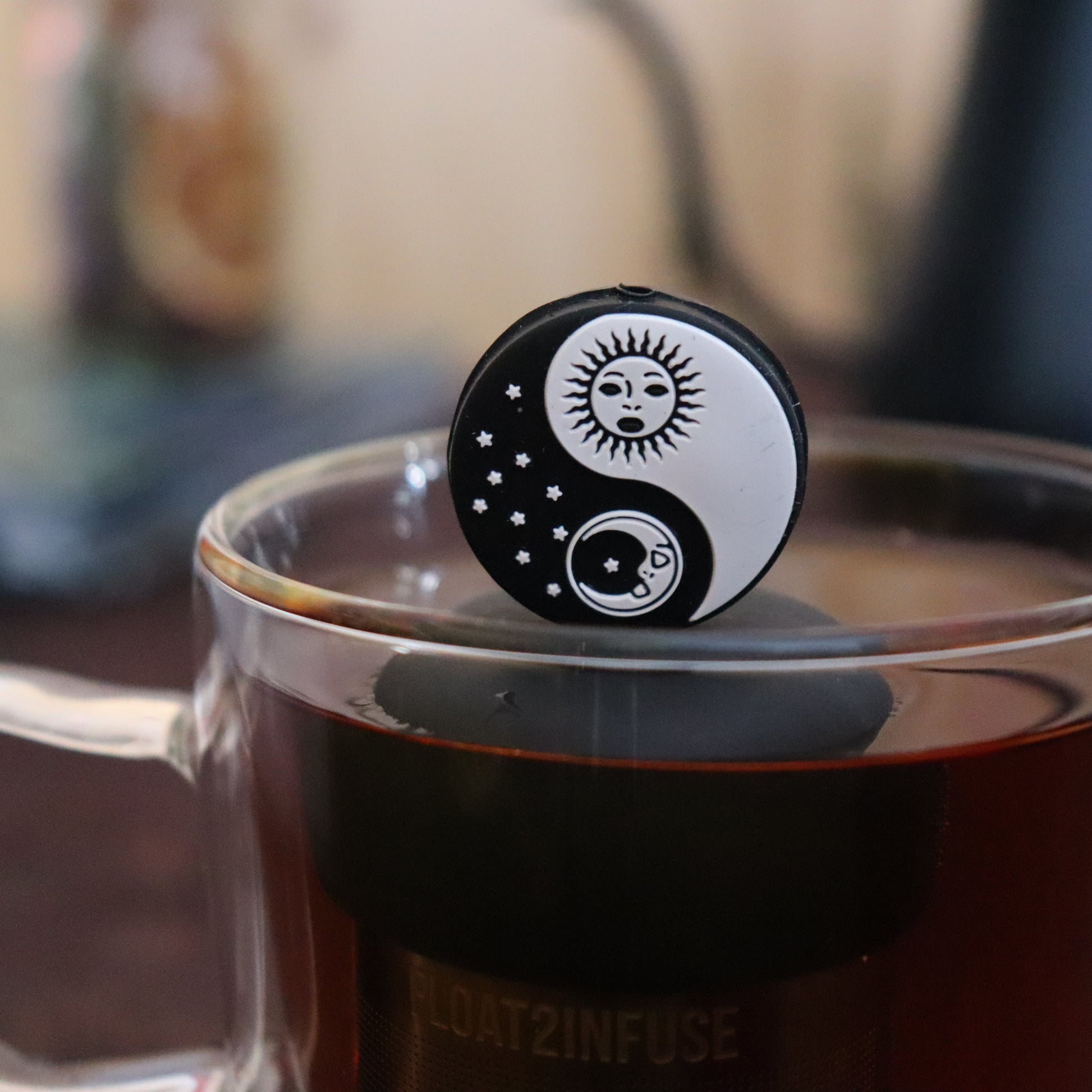 black and white sun and moon yin yang tea infuser for loose leaf tea  with a fine mesh stainless steel strainer that reads FLOAT2INFUSE on a black base floating in a glass tea mug with tea kettle in the background