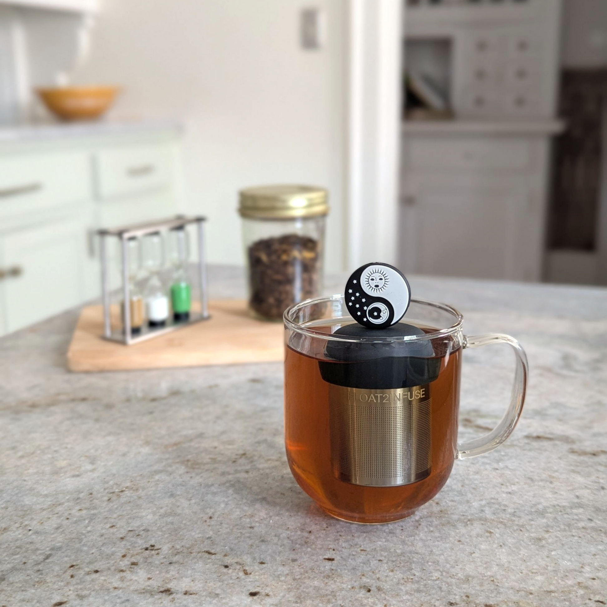 black and white yin yang with sun and moon tea infuser for loose leaf tea  with a fine mesh stainless steel tea infuser that reads FLOAT2INFUSE on a black base floating in a glass tea mug with loose leaf tea in the background