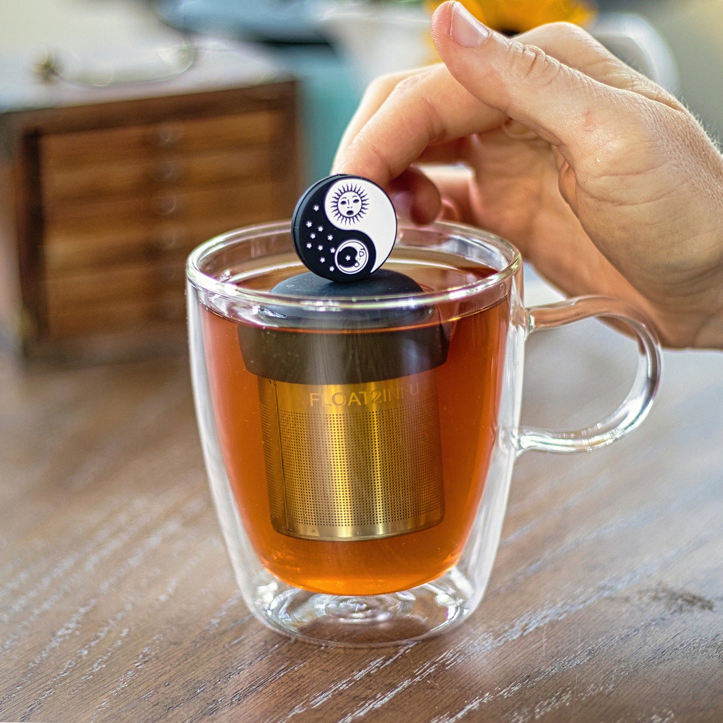 black and white sun and moon yin yang tea infuser for loose leaf tea  with a fine mesh stainless steel strainer that reads FLOAT2INFUSE on a black base floating in a glass tea mug with wooden jewelery box in the background