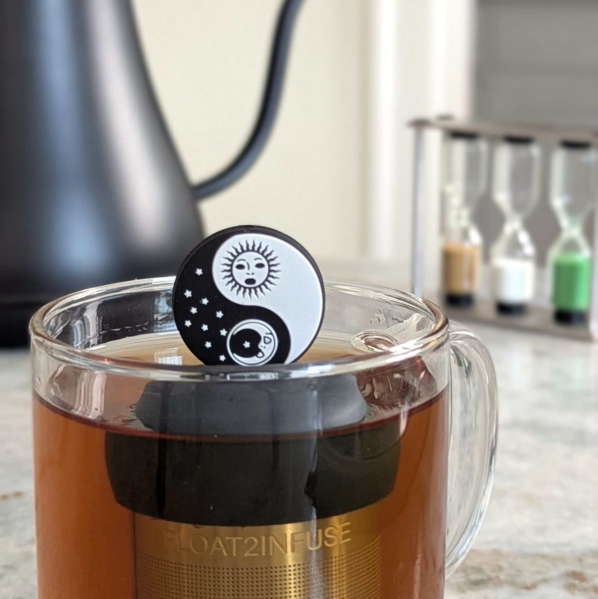 black and white sun and moon yin yang tea infuser for loose leaf tea  with a fine mesh stainless steel strainer that reads FLOAT2INFUSE on a black base floating in a glass tea mug with tea kettle in the background