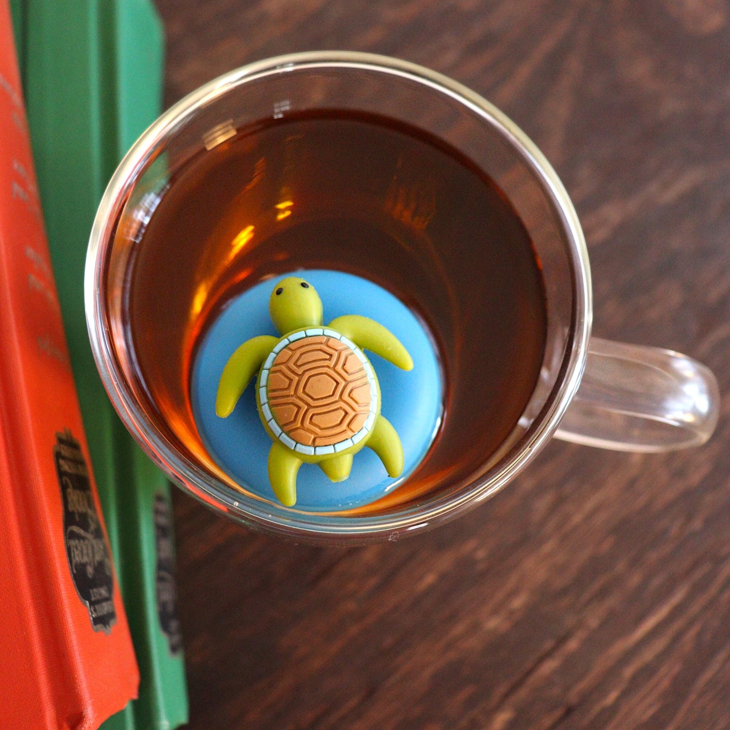 Aerial view of a cute green sea turtle with brown shell tea infuser for loose leaf tea with a fine mesh stainless steel tea strainer that reads FLOAT2INFUSE on a blue base floating in a glass tea mug with books to the side
