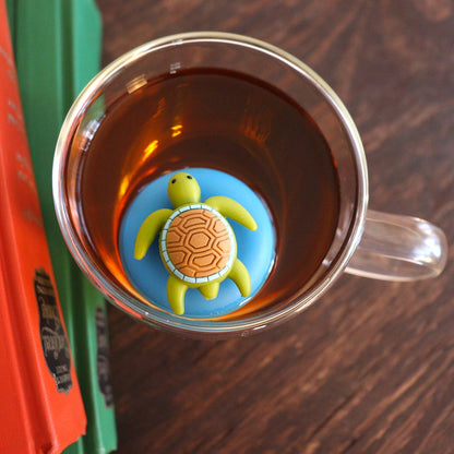 Aerial view of a cute green sea turtle with brown shell tea infuser for loose leaf tea with a fine mesh stainless steel tea strainer that reads FLOAT2INFUSE on a blue base floating in a glass tea mug with books to the side
