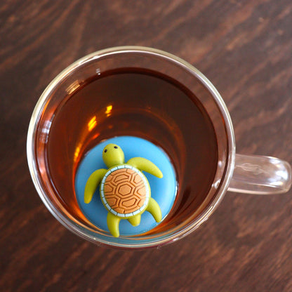 Aerial view of a cute green sea turtle with brown shell tea infuser for loose leaf tea with a fine mesh stainless steel tea strainer that reads FLOAT2INFUSE on a blue base floating in a glass tea mug
