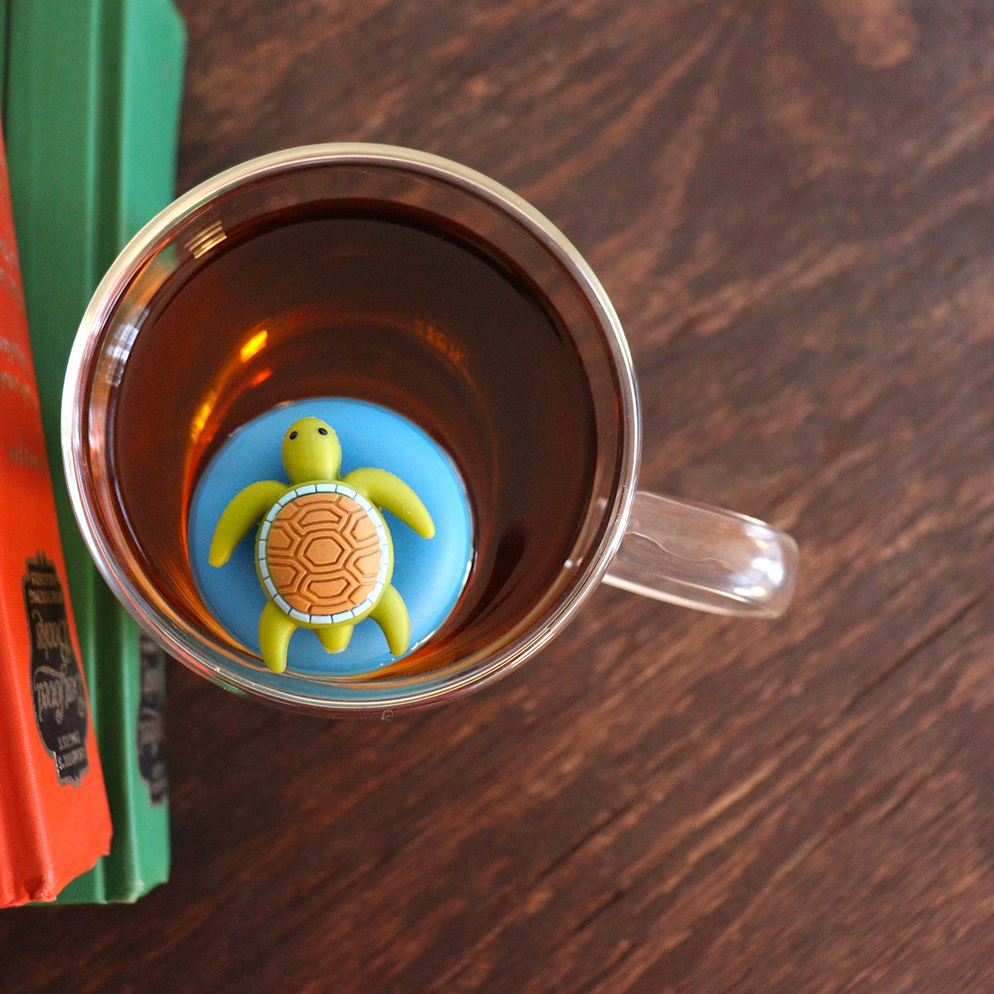 Aerial view of a cute green sea turtle with brown shell tea infuser for loose leaf tea with a fine mesh stainless steel tea strainer that reads FLOAT2INFUSE on a blue base floating in a glass tea mug with books to the side
