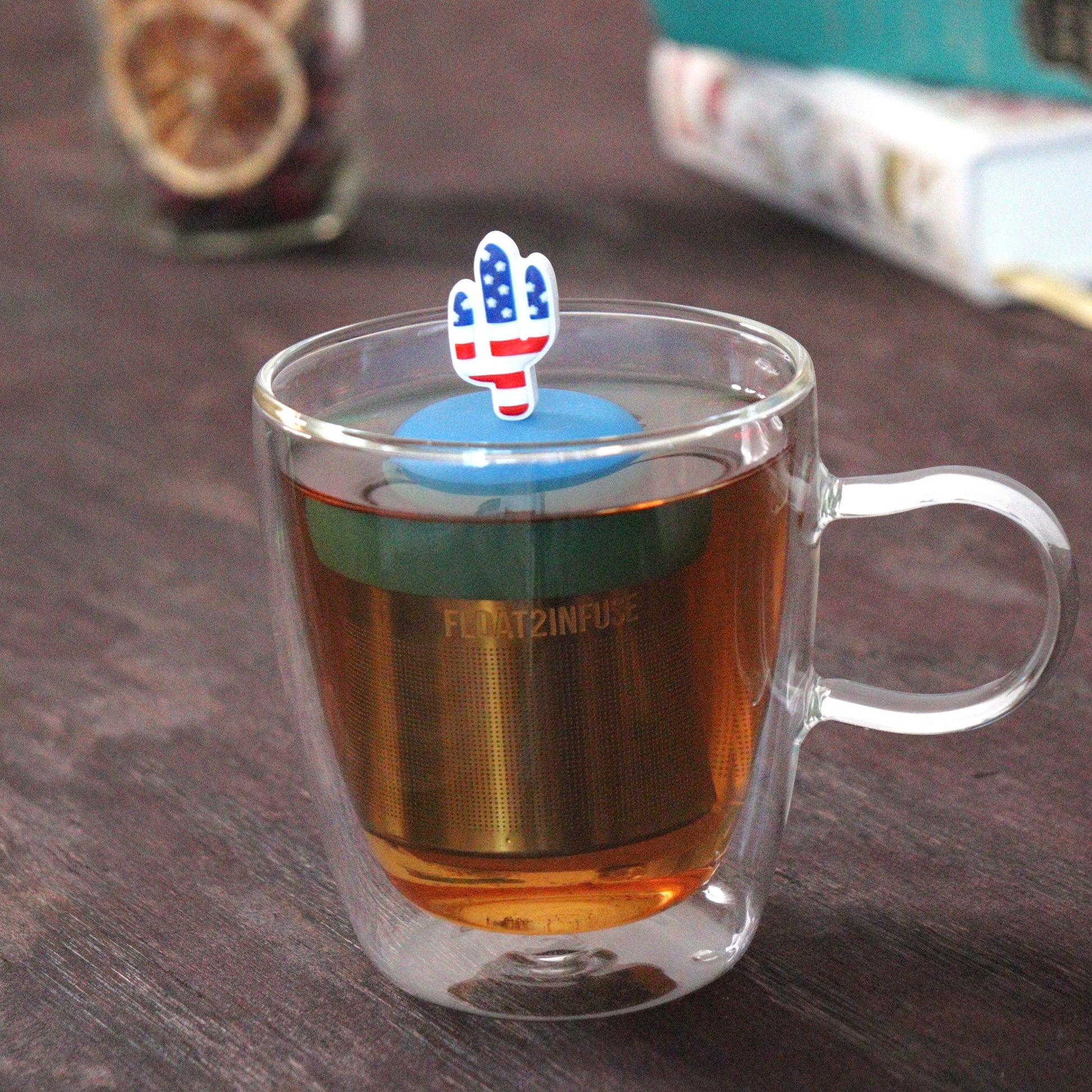American flag cactus tea infuser for loose leaf tea  with a fine mesh stainless steel tea strainer that reads FLOAT2INFUSE on a blue base floating in a glass tea mug with loose leaf tea and books in the background