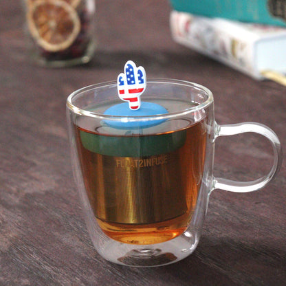 American flag cactus tea infuser for loose leaf tea  with a fine mesh stainless steel tea strainer that reads FLOAT2INFUSE on a blue base floating in a glass tea mug with loose leaf tea and books in the background