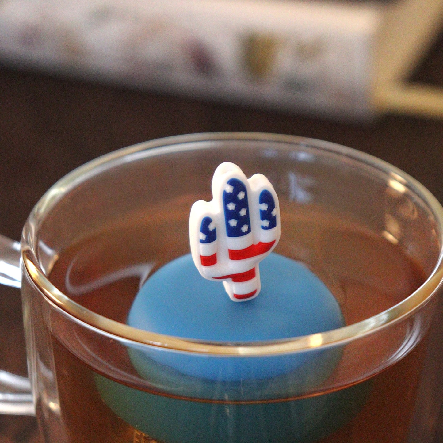 American flag cactus tea infuser for loose leaf tea  with a fine mesh stainless steel tea strainer that reads FLOAT2INFUSE on a blue base floating in a glass tea mug with books in the background