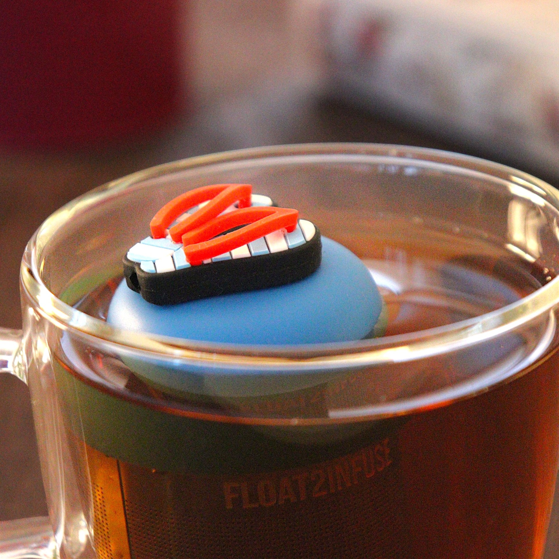 Americana blue and white striped flip flops with red straps tea infuser for loose leaf tea with a fine mesh stainless steel tea strainer that reads FLOAT2INFUSE on a blue base  floating in a glass tea mug 
with books in the background
