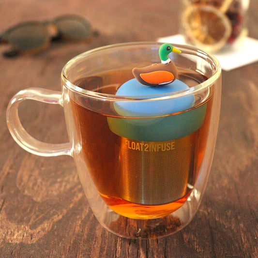 mallard duck tea infuser for loose leaf tea with a fine mesh stainless steel tea infuser that reads FLOAT2INFUSE on a blue base  floating in a glass tea mug with  loose leaf tea and sunglasses in the background