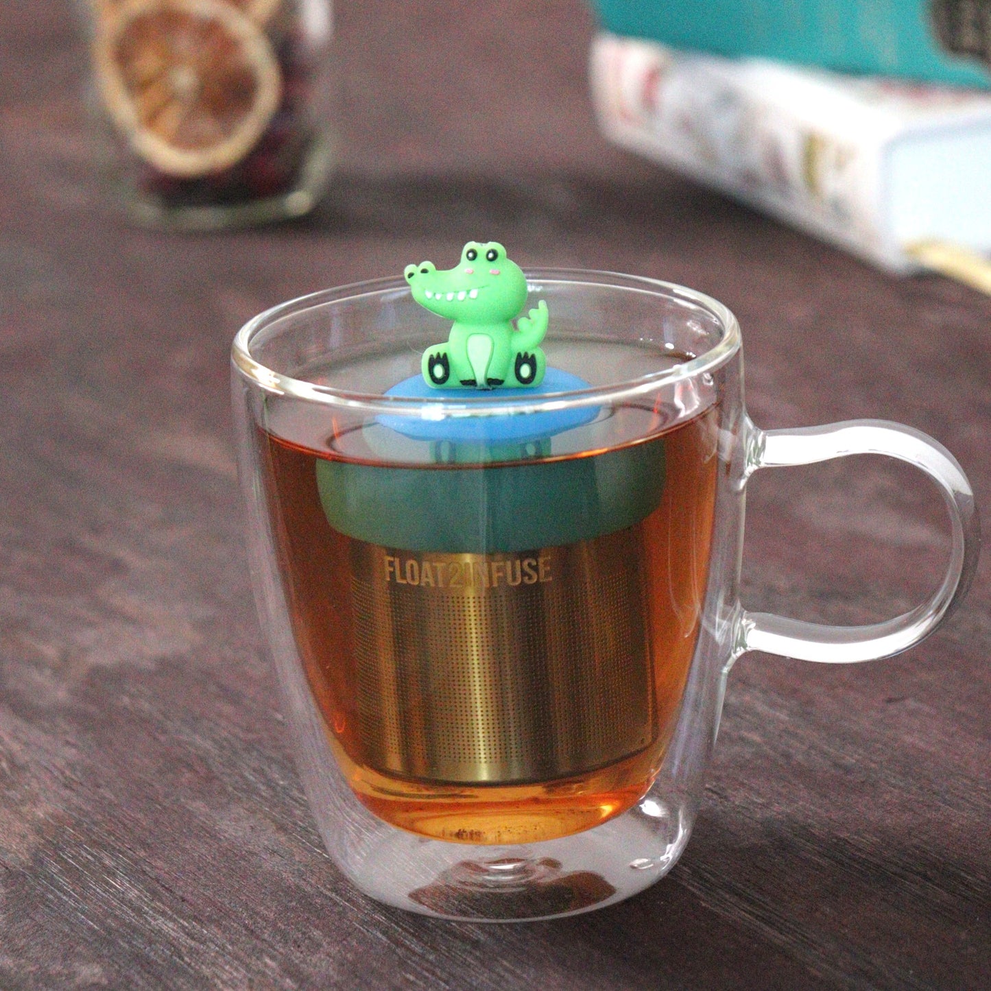 green alligator animal tea infuser for loose leaf tea with a fine mesh stainless steel tea strainer that reads FLOAT2INFUSE on a blue base floating in a glass tea mug with books in the background