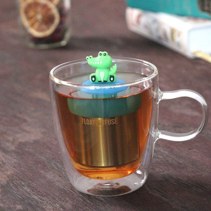 green alligator animal tea infuser for loose leaf tea with a fine mesh stainless steel tea strainer that reads FLOAT2INFUSE on a blue base floating in a glass tea mug with books in the background