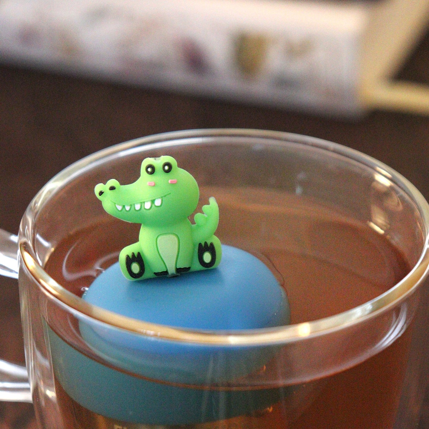 green alligator animal tea infuser for loose leaf tea  with a fine mesh stainless steel tea strainer that reads FLOAT2INFUSE on a blue base floating in a glass tea mug with books in the background