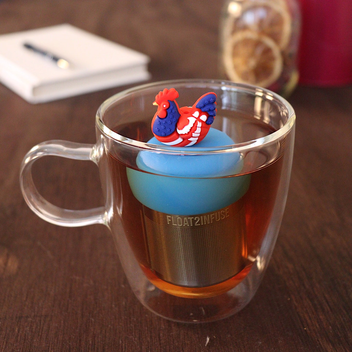 Red, white, and blue patterned chicken tea infuser for loose leaf tea with a fine mesh stainless steel tea strainer 
that reads FLOAT2INFUSE on a blue base with a pen laying across a book and dried orange slices in a mason jar in the background
