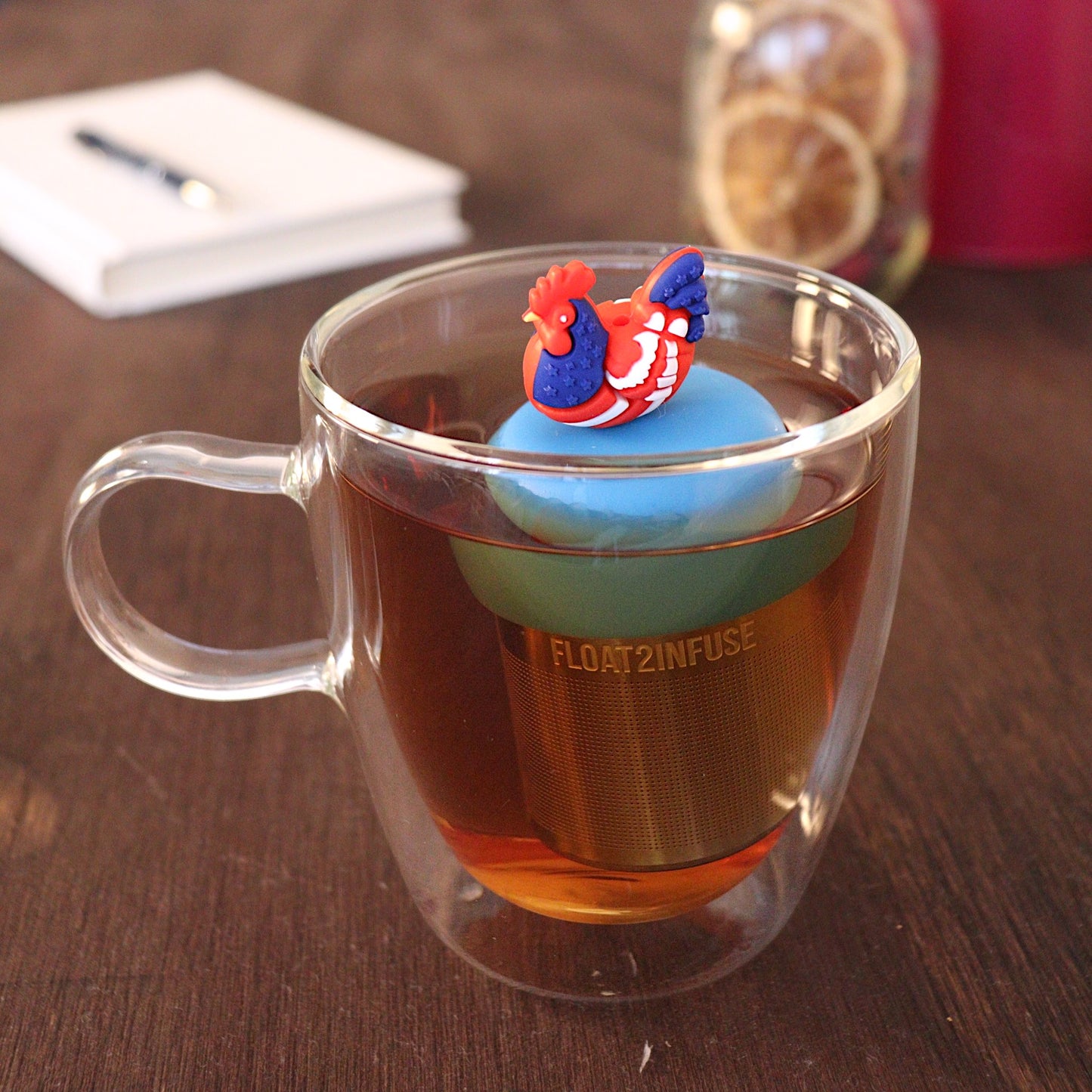 Red, white, and blue patterned chicken tea infuser for loose leaf tea with a fine mesh stainless steel tea strainer 
that reads FLOAT2INFUSE on a blue base with a pen laying across a book and dried orange slices in a mason jar in the background