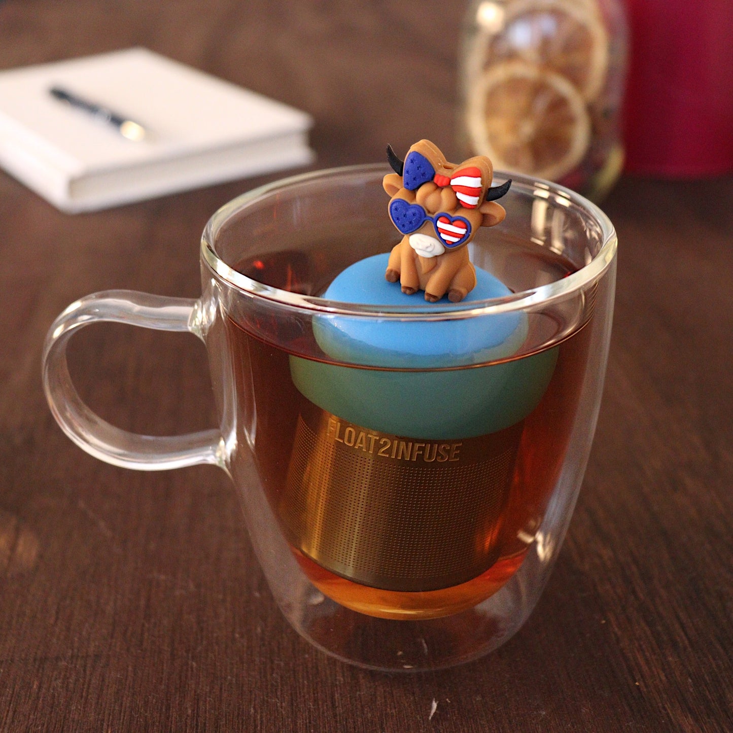 Cute cow wearing red white and blue heart shaped glasses and hairbow tea infuser for loose leaf tea with a fine mesh stainless steel tea strainer that reads FLOAT2INFUSE on a blue base with a pen laying across a book and dried orange slices in a mason jar in th background