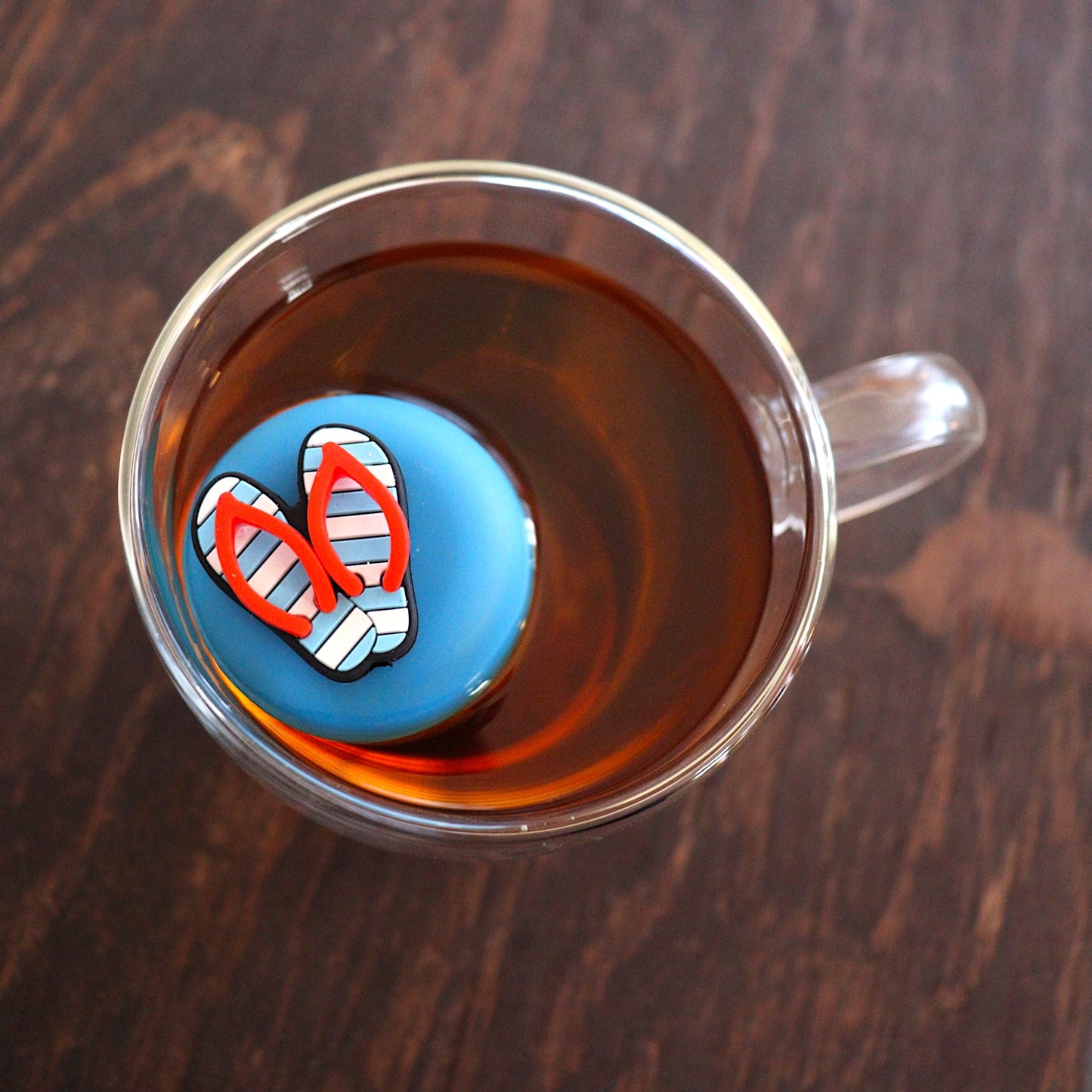 Aerial view of Americana blue and white striped flip flops with red straps tea infuser for loose leaf tea with a fine mesh stainless steel tea strainer that reads FLOAT2INFUSE on a blue base  floating in a glass tea mug 