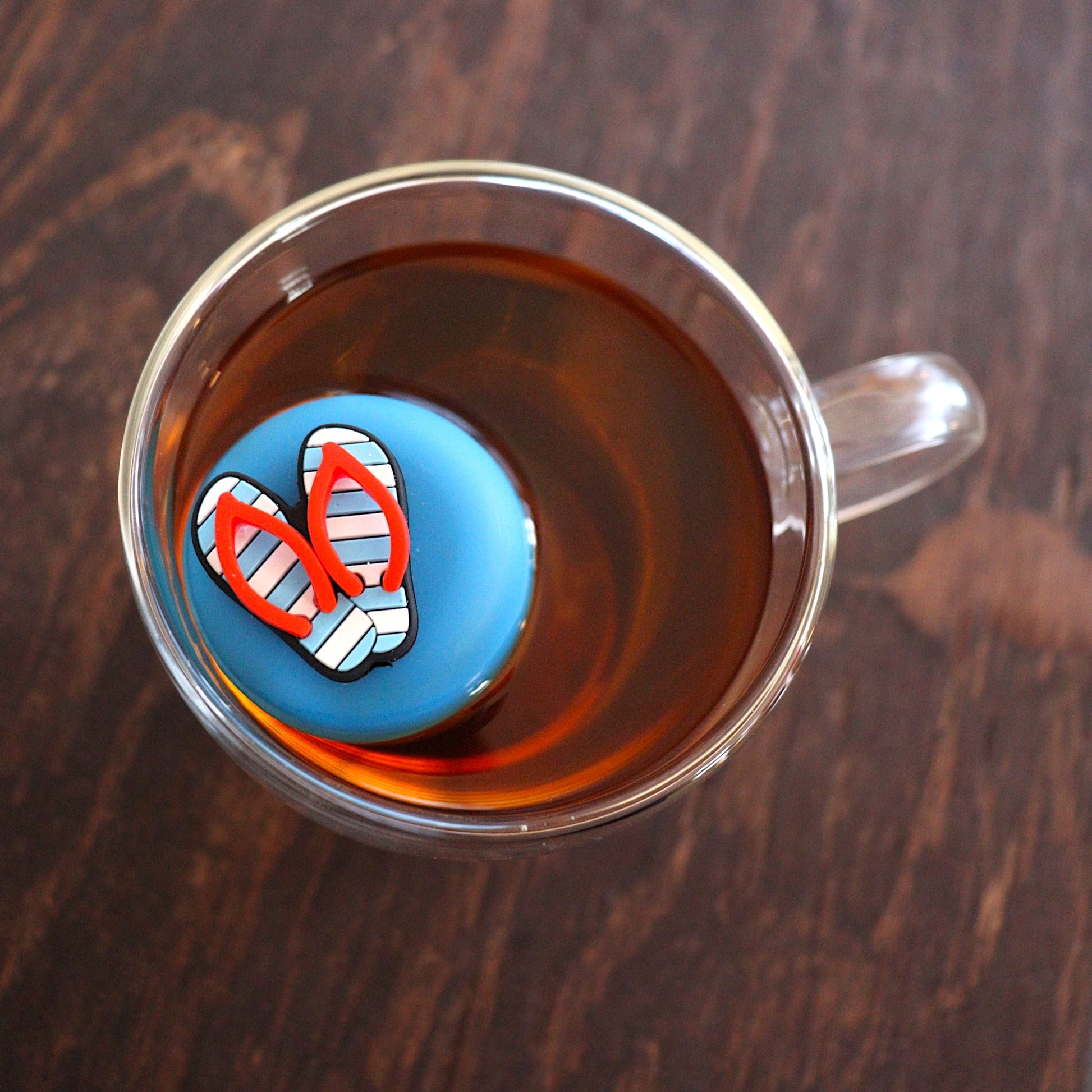 Aerial view of Americana blue and white striped flip flops with red straps tea infuser for loose leaf tea with a fine mesh stainless steel tea strainer that reads FLOAT2INFUSE on a blue base  floating in a glass tea mug 