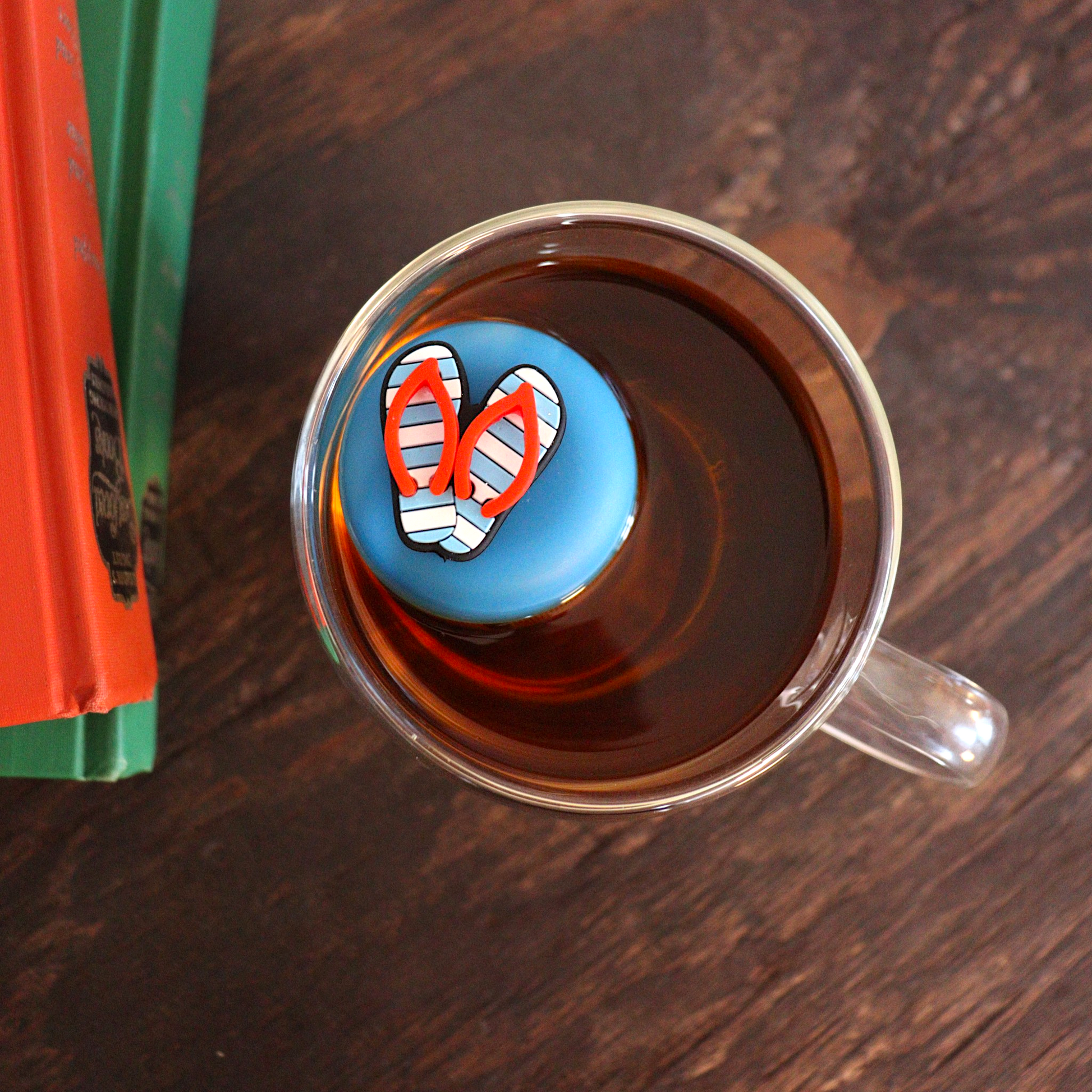 Aerial view of Americana blue and white striped flip flops with red straps tea infuser for loose leaf tea with a fine mesh stainless steel tea strainer that reads FLOAT2INFUSE on a blue base  floating in a glass tea mug with books in the background