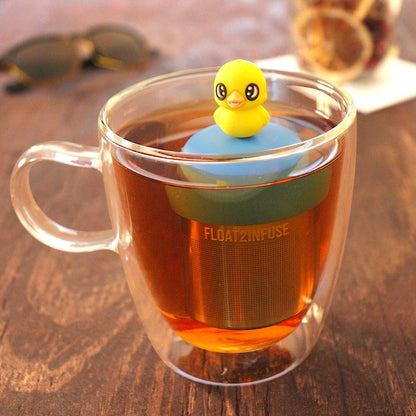 Baby duck animal tea infuser for loose leaf tea with a fine mesh stainless steel tea infuser that reads FLOAT2INFUSE on a blue base  floating in a glass tea mug with  loose leaf tea and sunglasses in the background