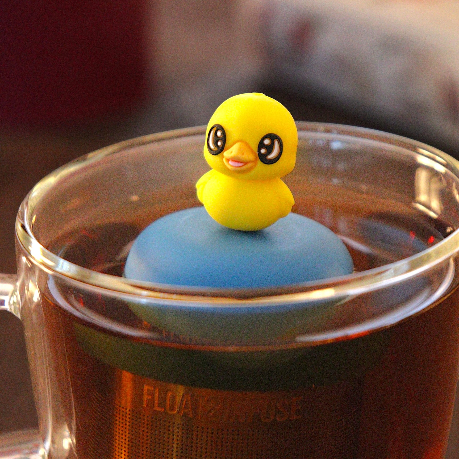 Baby duck animal tea infuser for loose leaf tea with a fine mesh stainless steel tea infuser that reads FLOAT2INFUSE on a blue base  floating in a glass tea mug with  loose leaf tea and books in the background