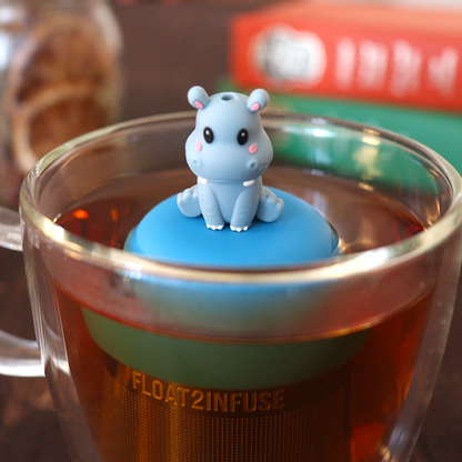 grey smiling baby hippo animal tea infuser for loose leaf tea  with a fine mesh stainless steel tea infuser that reads FLOAT2INFUSE on a blue base floating in a glass tea mug with loose leaf tea and books in the background