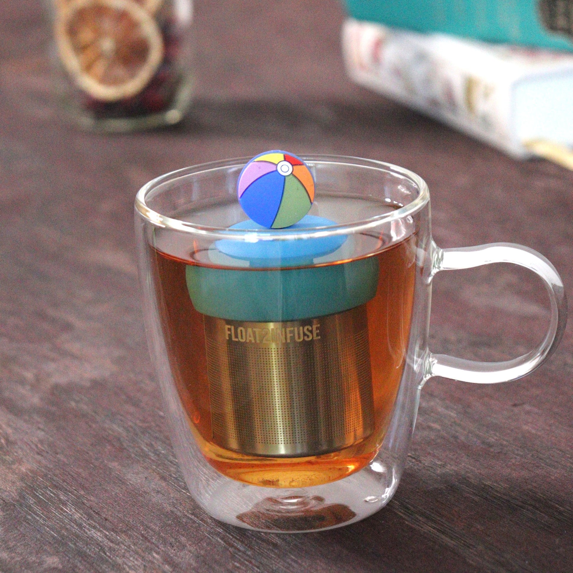 beach ball tea infuser for loose leaf tea with a fine mesh stainless steel tea strainer that reads FLOAT2INFUSE on a blue base floating in a glass tea mug with loose leaf tea and books in the background