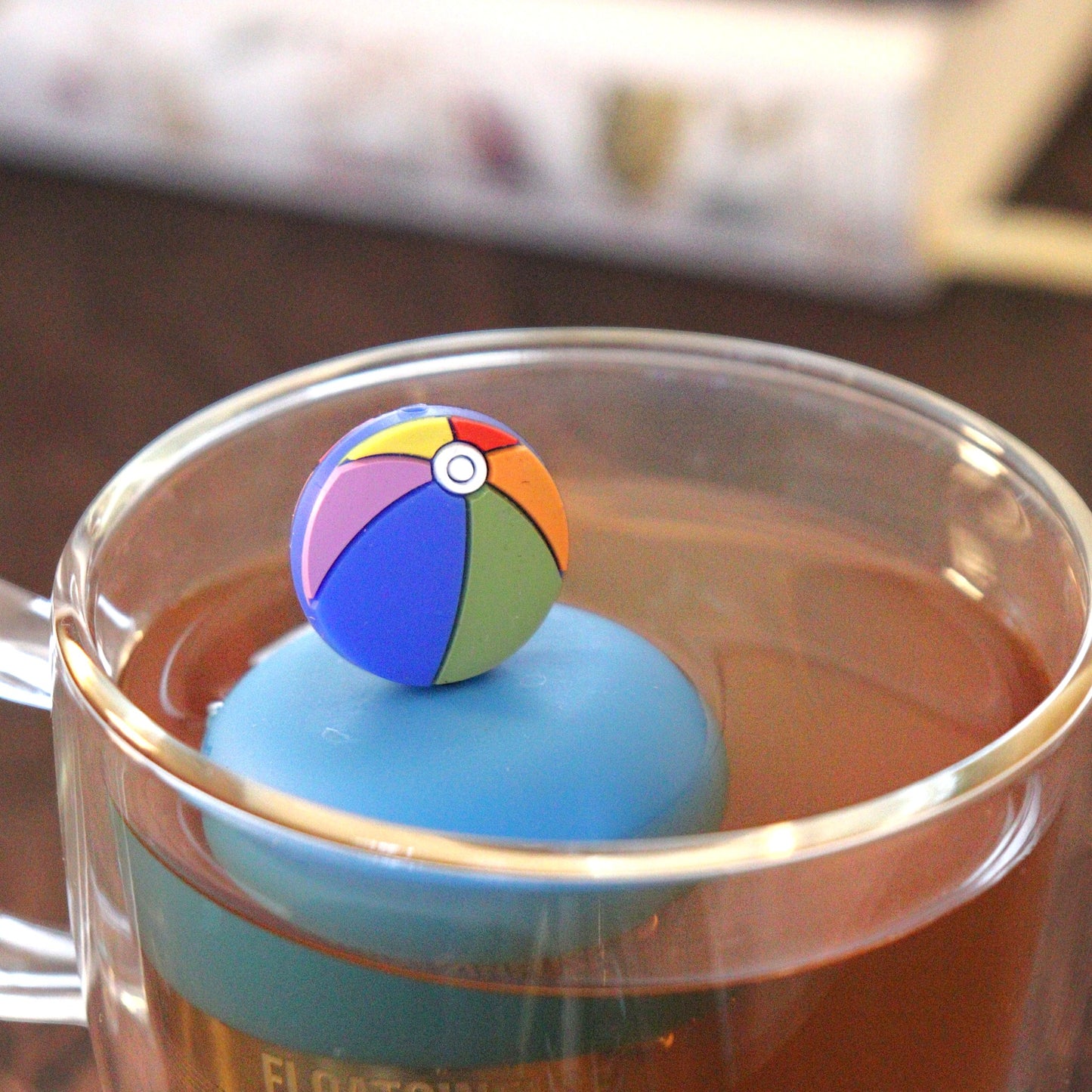 beach ball tea infuser for loose leaf tea e with a fine mesh stainless steel tea strainer that reads FLOAT2INFUSE on a blue bas floating in a glass tea mug with books in the background